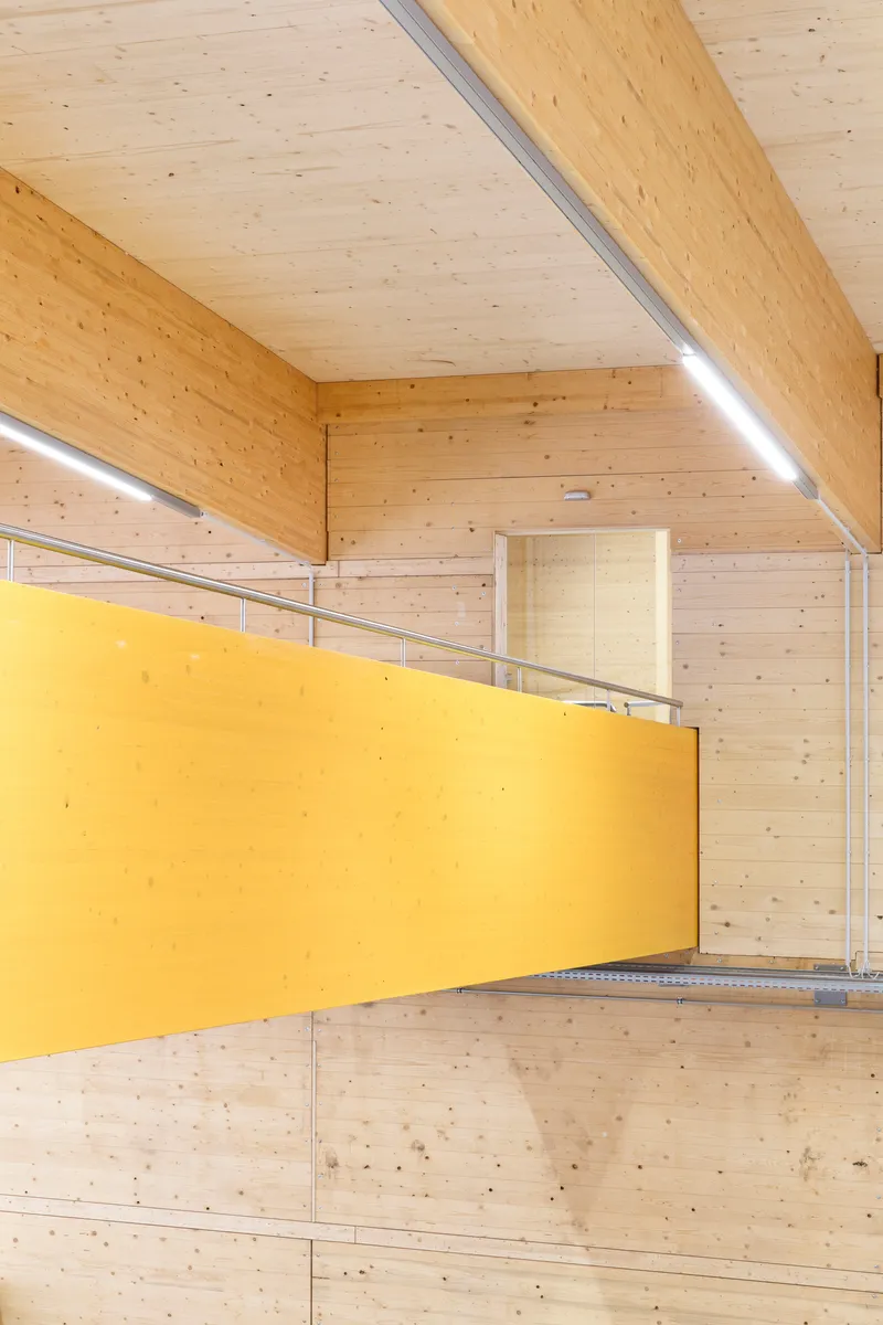 Yellow mezzanine walkway with metal railing suspended within double-height space clad in exposed timber planks