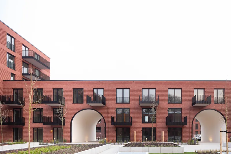 Red brick facade with recessed balconies and twin arched passageways flanked by young planted trees