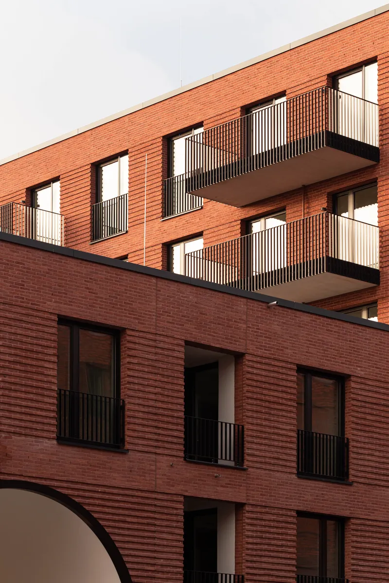 Corner detail of the red brick facade showing stacked balconies with vertical louvered screens in afternoon light