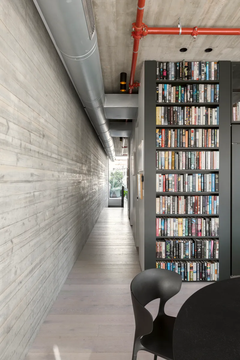 Narrow corridor with board-formed concrete wall, exposed ductwork, and tall bookshelf along the passage