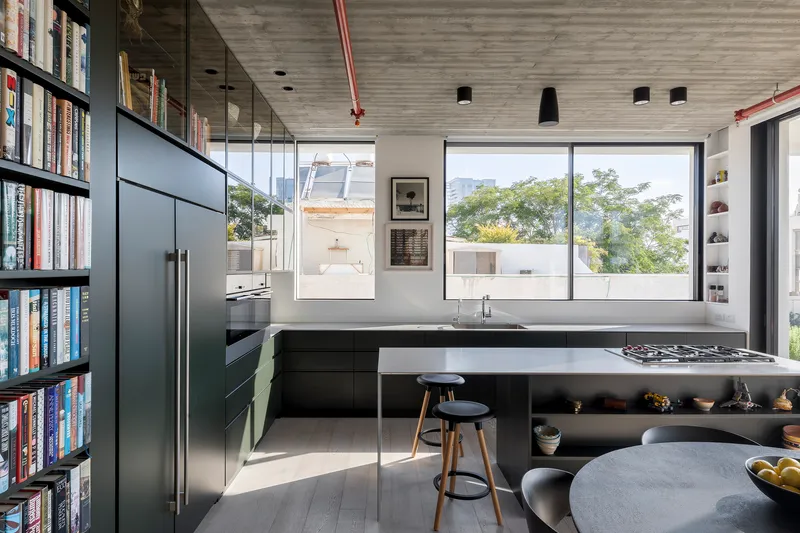 Kitchen island with integrated cooktop beside full-height glazed partition reflecting trees outside in afternoon light