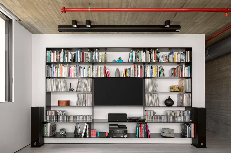 Built-in black shelving unit with television and books below exposed concrete ceiling with red conduit
