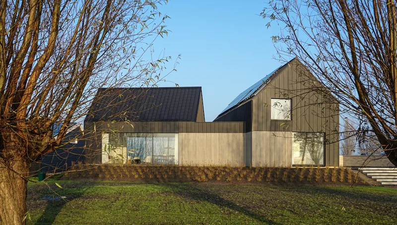 Two gabled volumes clad in light timber and dark metal standing behind bare winter trees at dusk
