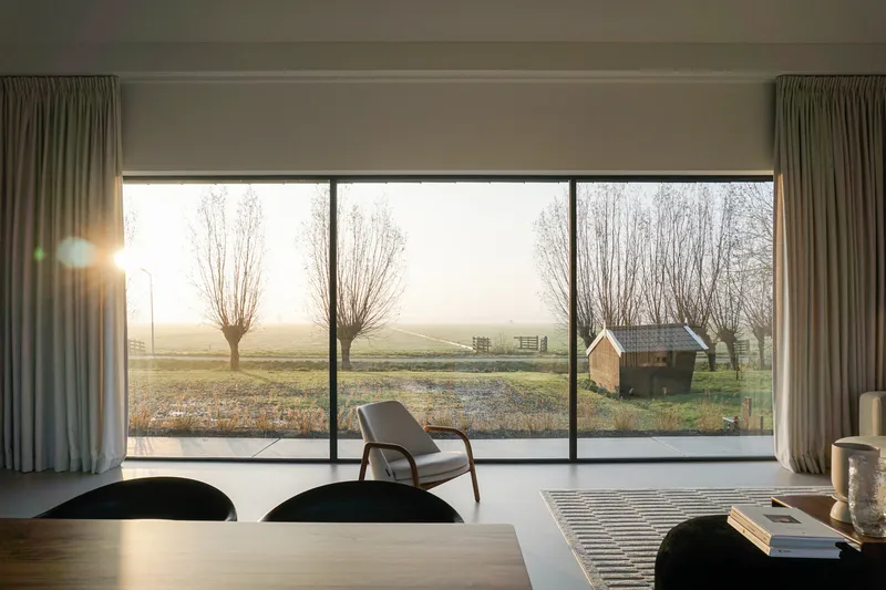 Interior view through floor-to-ceiling glazing overlooking a misty pasture with pollarded willows at dawn