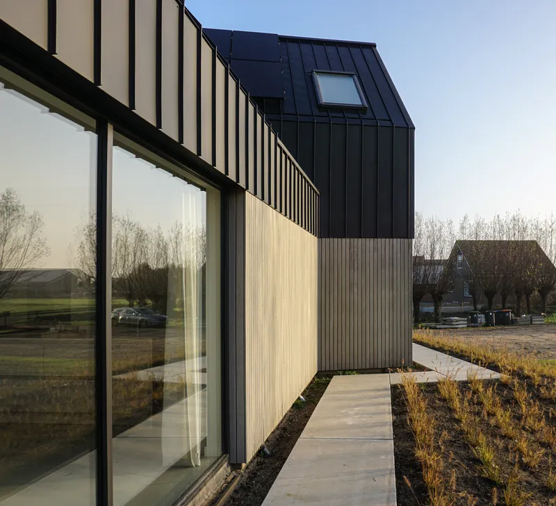 Concrete walkway alongside dark metal and timber facade with planted beds of dried grasses
