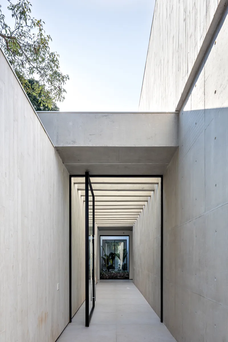 Narrow concrete passageway with open skylight and rhythmic timber beam shadows leading to a glazed door
