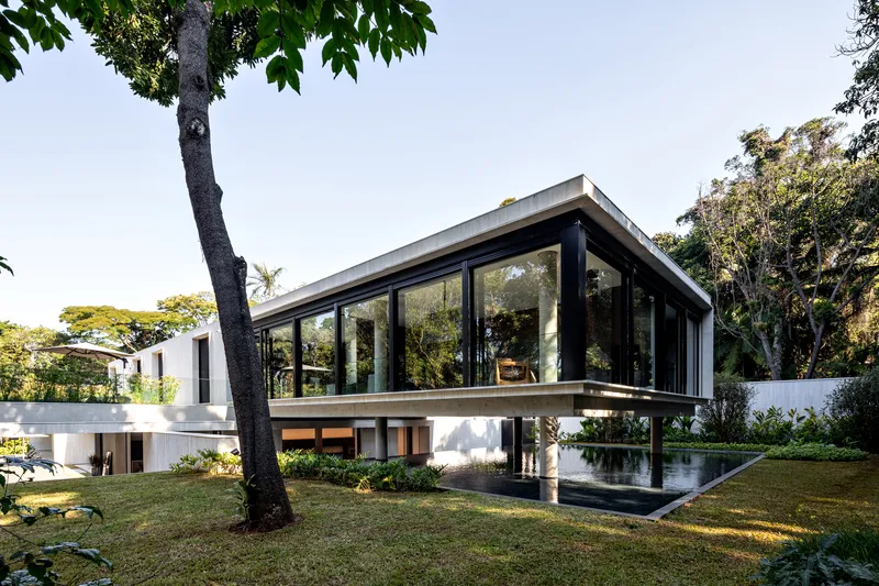 Glazed upper pavilion on concrete columns beside a reflecting pool and lawn framed by trees