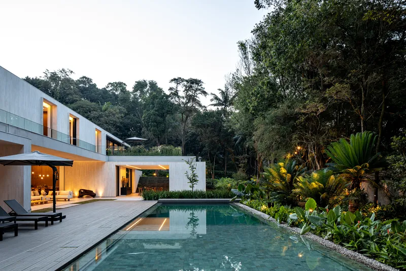 Terrace view of the pool and illuminated interior spaces at dusk backed by dense forest
