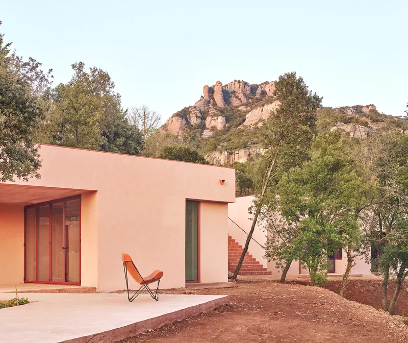 Pink stucco facade with timber-framed glazing and an orange butterfly chair beneath rocky hillside vegetation