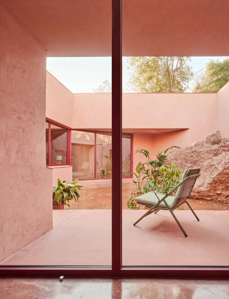 Interior view through floor-to-ceiling glazing onto a pink courtyard with chair and potted palms