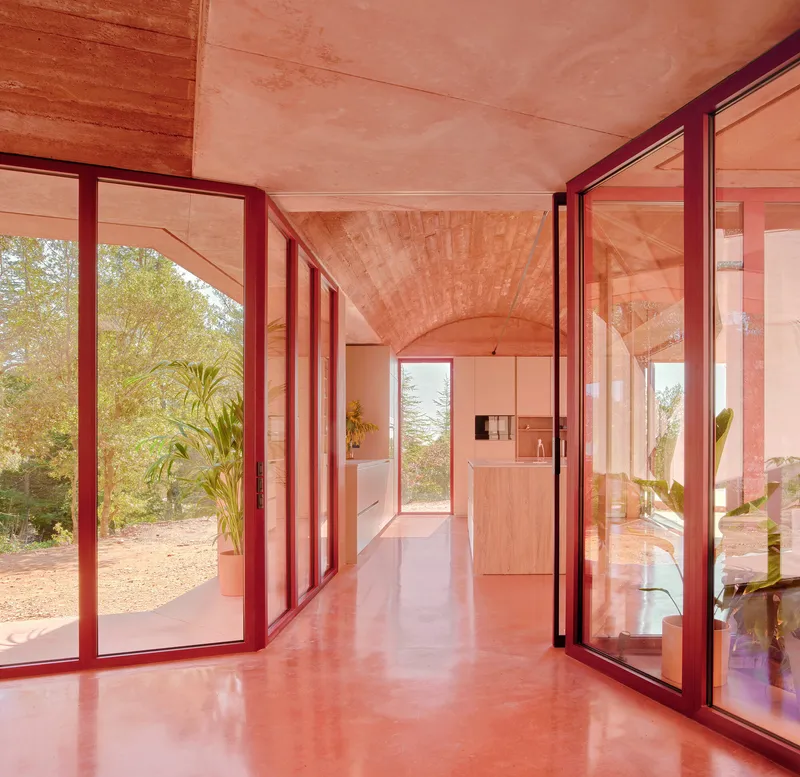 Corridor with polished pink concrete floor, red metal-framed glazing, and vaulted timber ceiling in autumn light