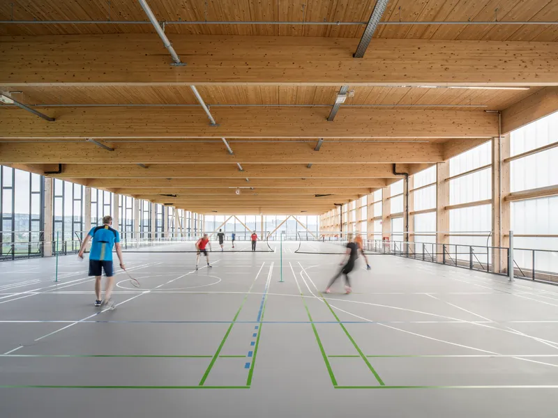 Interior sports hall with exposed timber beams and glazed sidewalls with people playing badminton