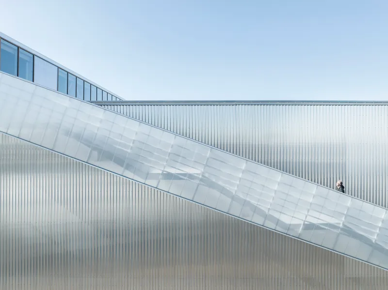 Diagonal skybridge with translucent paneling and a lone figure ascending the ramp
