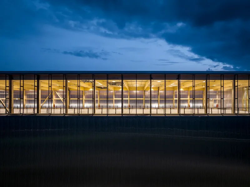 Continuous glass facade with timber ceiling visible inside under dark evening sky