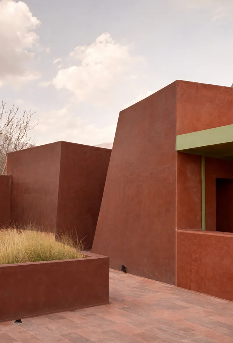 Exterior corner showing red stucco volumes and a planted bed under cloudy skies
