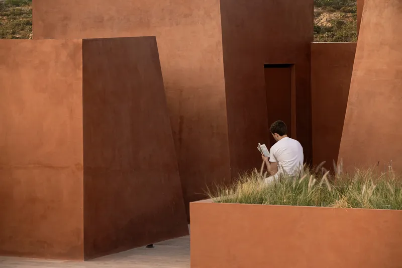 Visitor seated reading beside a pivoting steel door and planted grasses in daylight