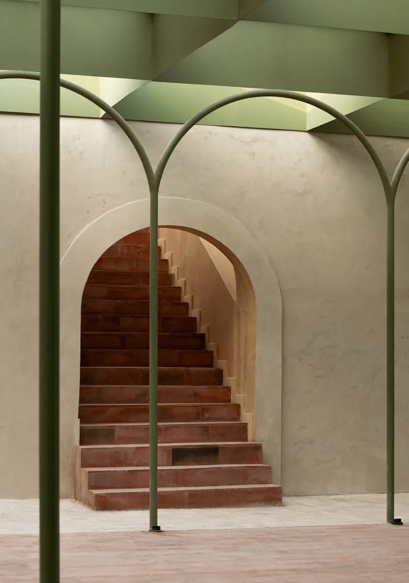 View through green steel arch columns toward the curved terracotta staircase in its vaulted alcove