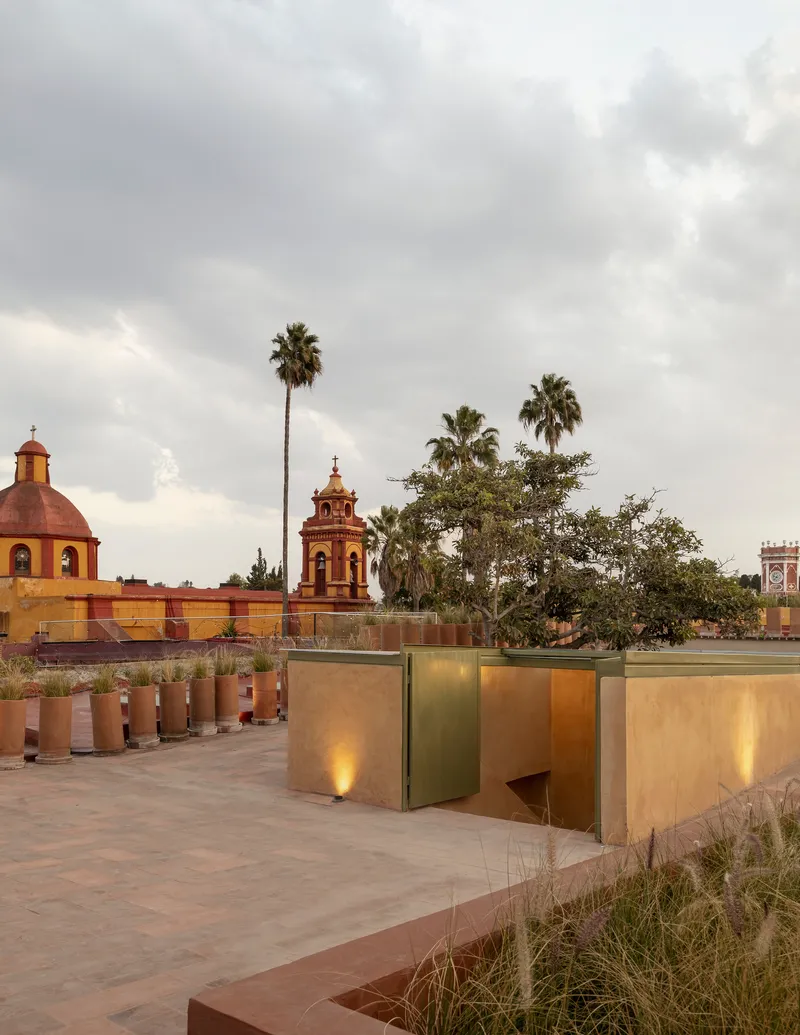 Rooftop terrace with brass-clad volumes and planted beds under cloudy skies with palms and church towers beyond