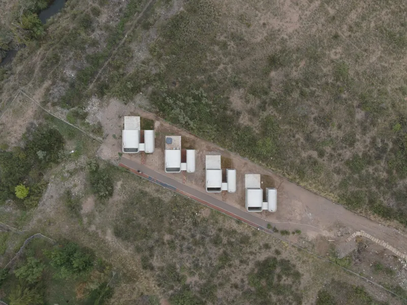 Drone view of paired white volumes arranged in a row beside a gravel road through sparse vegetation