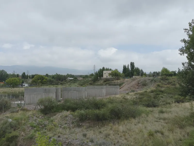 Board-formed concrete retaining wall set into a hillside with scrubland vegetation under overcast sky