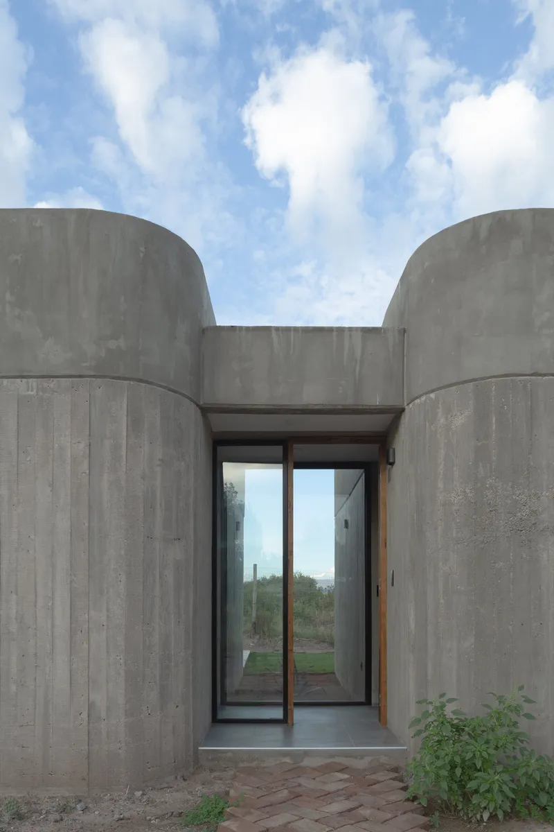 Glass entry door set between two curving board-formed concrete volumes under a blue sky