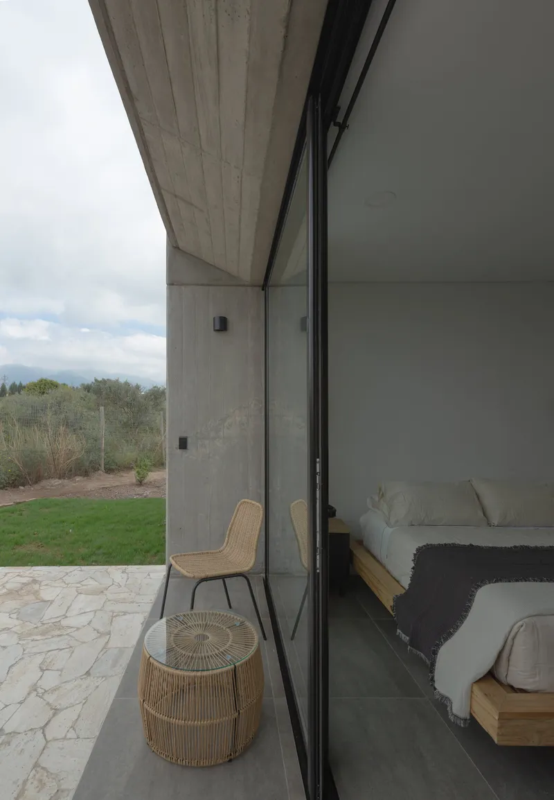 Sliding glass wall opening from bedroom to paved terrace with woven furniture overlooking scrubland