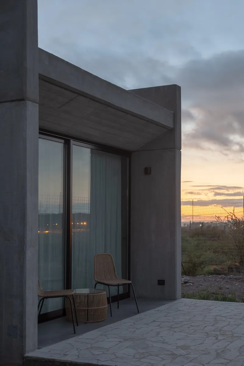 Concrete overhang above floor-to-ceiling glazing with wooden chairs on a stone terrace at sunset