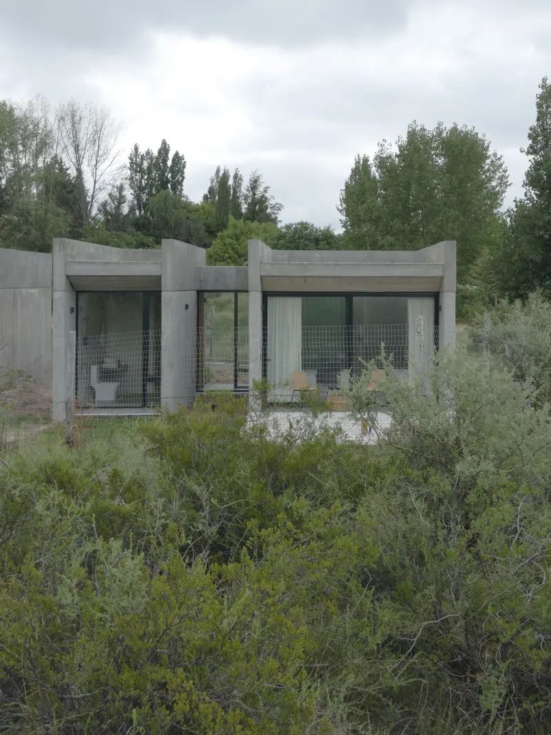 Two-lobed concrete structure with glazed openings surrounded by dense scrubland under cloudy skies