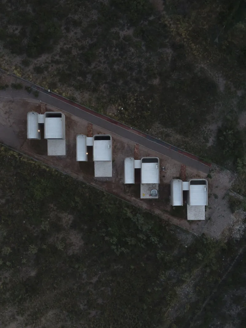 Aerial view of four white rectangular housing units aligned along a roadway in scrubland at dusk