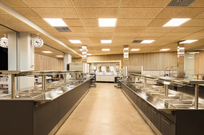Commercial kitchen with stainless steel counters and glass sneeze guards beneath linear suspended light fixtures