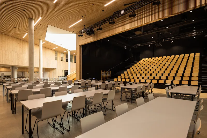 Multipurpose hall with retractable seating, white folding tables and exposed slatted timber ceiling with skylights