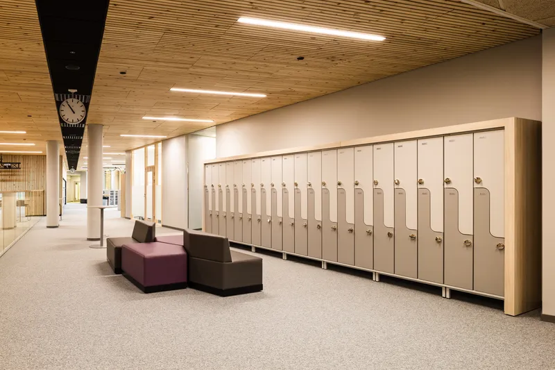 Corridor with pale locker bank and modular soft seating beneath timber acoustic ceiling and linear lighting
