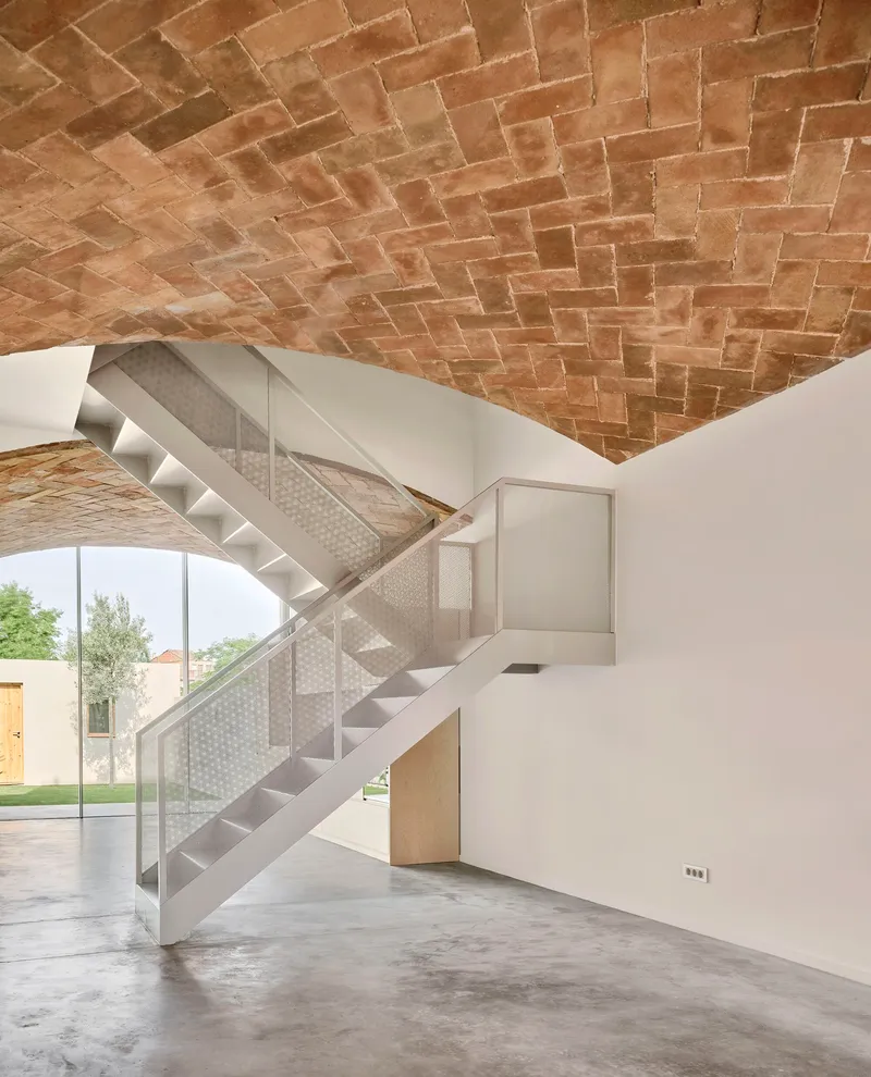 Perforated metal staircase beneath a herringbone brick vault with garden visible through glazed opening