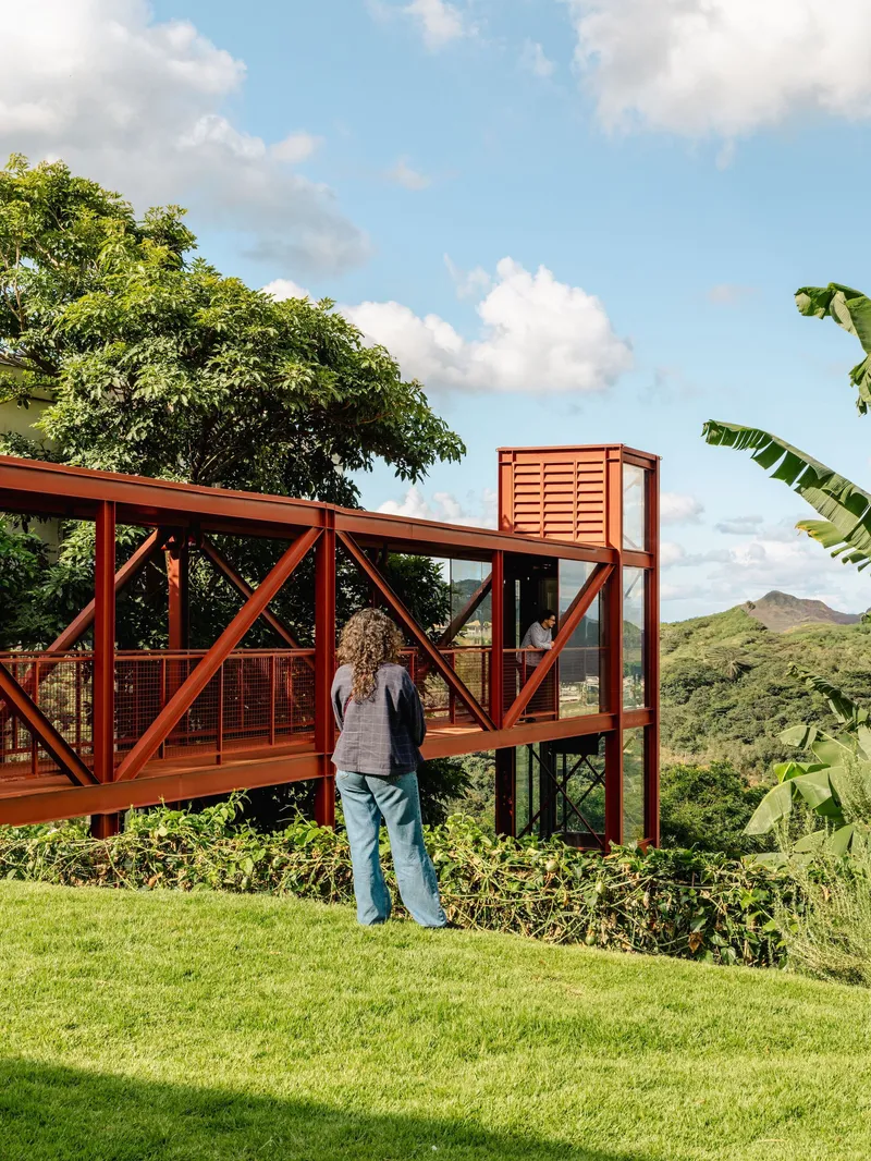 Red steel frame bridge structure with cross-bracing extending over hillside vegetation as a person looks out