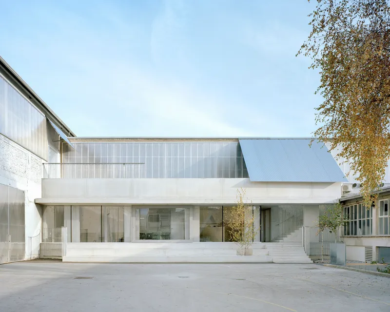 White concrete and translucent polycarbonate facade with cantilevered upper floor and young tree in courtyard
