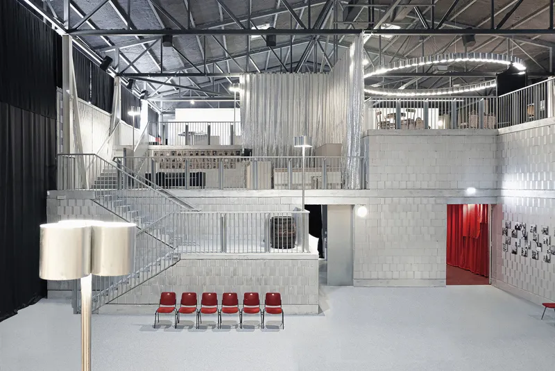 Triple-height interior hall with exposed steel trusses, metal railings, concrete platforms and row of red chairs