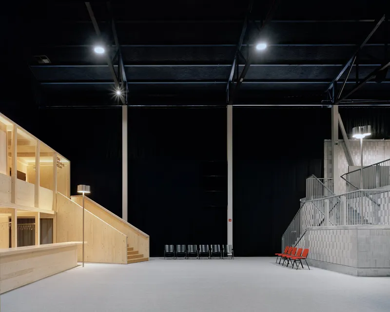 Black-painted ceiling with exposed steel structure above white resin floor and plywood volume in darkness