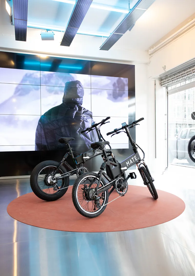 Three bicycles on a circular terracotta platform beneath a video wall showing a figure