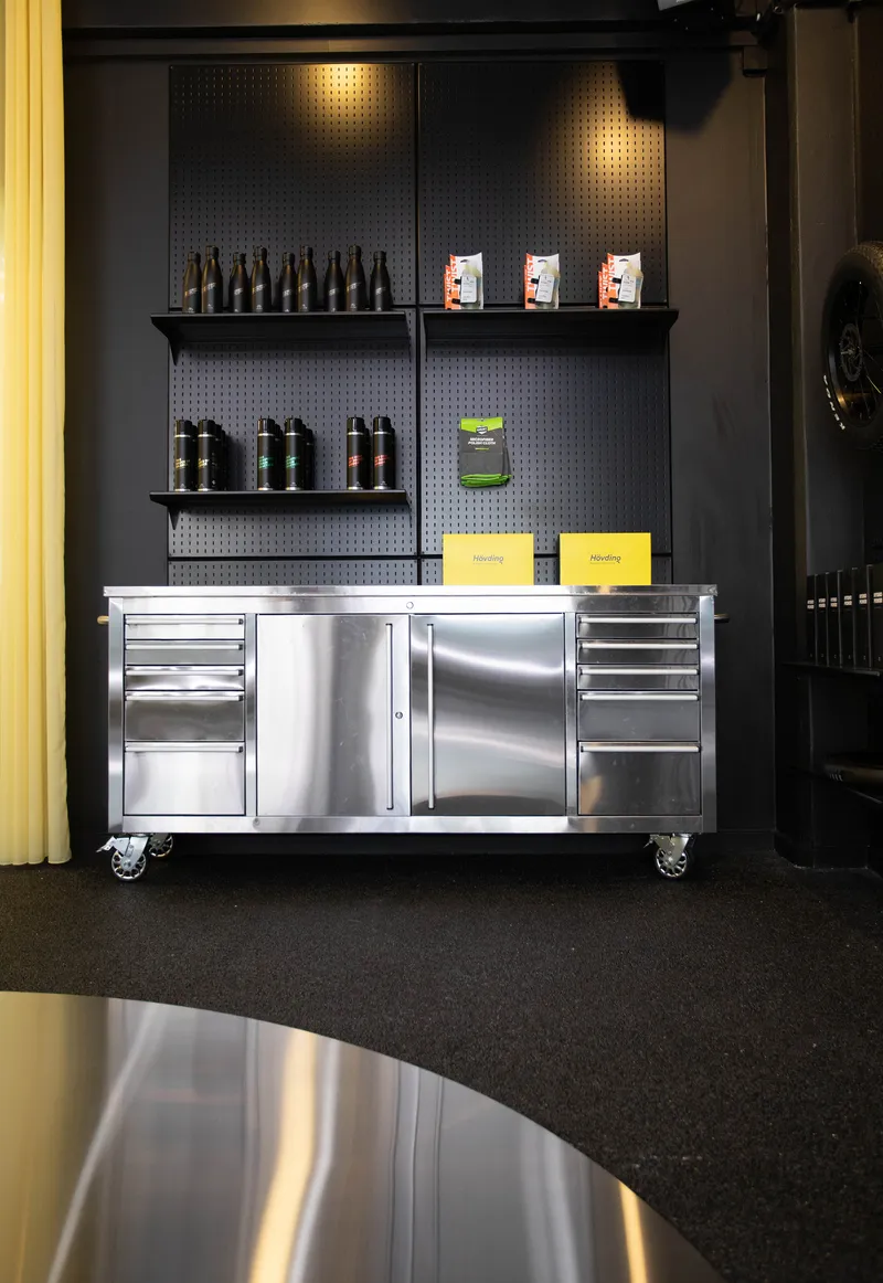 Stainless steel rolling cart with storage drawers below a black pegboard wall with shelving and yellow curtain