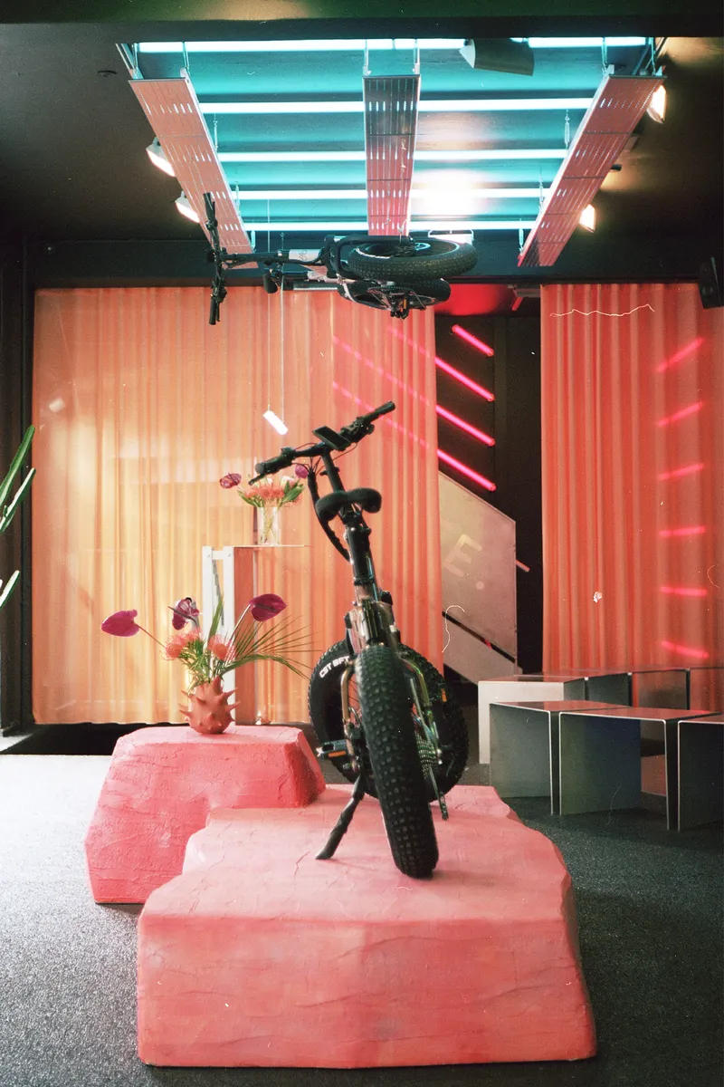 Bicycle displayed on pink foam blocks beneath ceiling-mounted bike and multicolored neon lighting