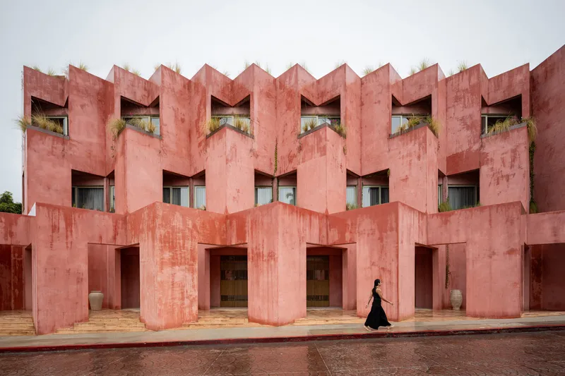 Zigzag facade of pink plastered concrete with deep window recesses and a figure in black passing below