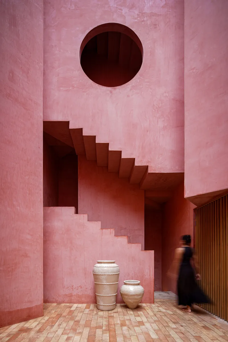 Interior staircase rising beneath a circular void with ceramic vessels at its base and a passing figure