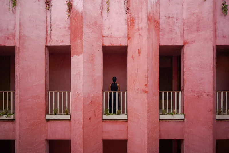 Facade detail with vertical pink concrete fins and a figure standing on a balcony with striped railings