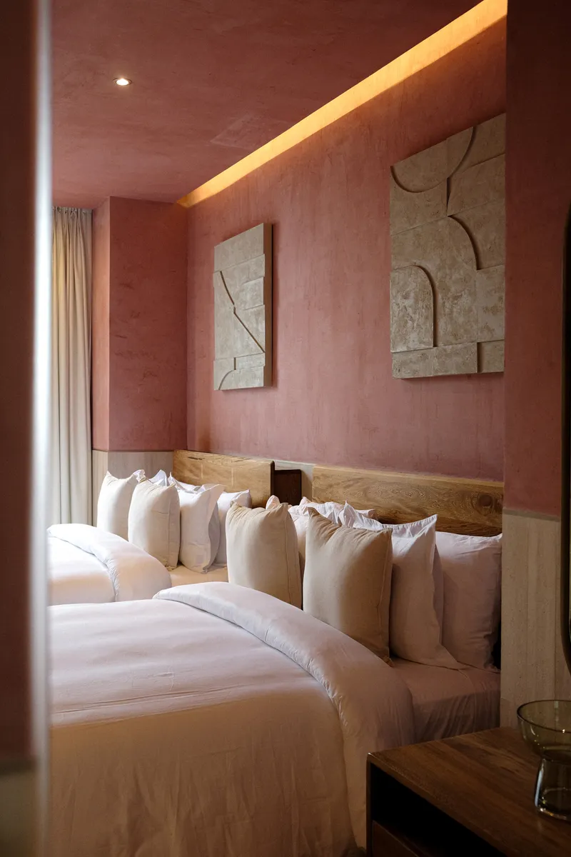 Bedroom with pink plaster walls, timber headboard, white linens and stone relief panels with warm cove lighting