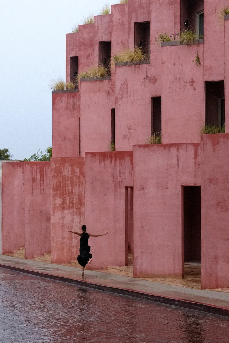 Tiered red plaster volumes with planted openings as a figure stands with outstretched arms on wet pavement