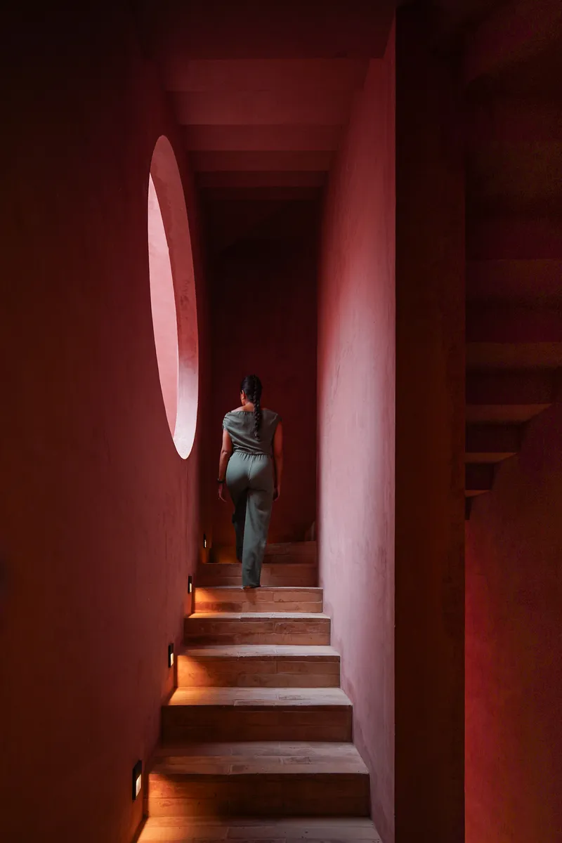 Person ascending a narrow red plastered staircase with oval wall opening and recessed step lighting