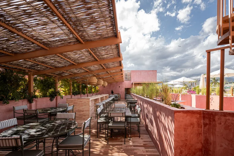 Rooftop terrace with woven timber pergola, dining tables and pink plaster volumes under cloudy sky