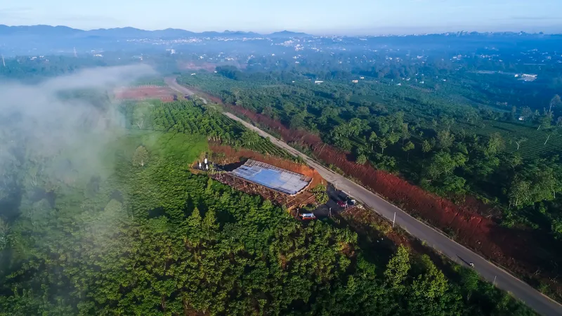 Aerial view of a low-slung roof nestled among agricultural plots and forested hillsides in morning mist