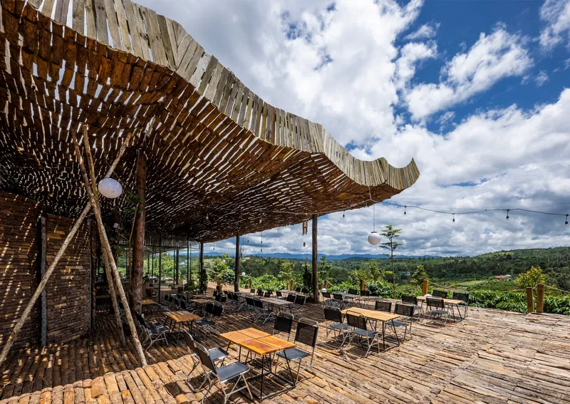 Open-air terrace with woven bamboo canopy and timber columns overlooking a forested valley under scattered clouds