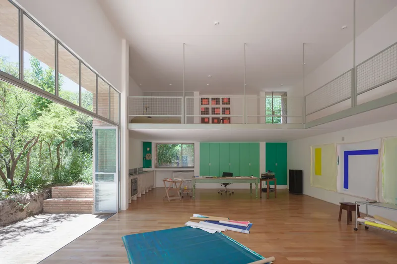 Double-height studio space with timber floors, wire mesh mezzanine railing, and colorful geometric canvases on white walls